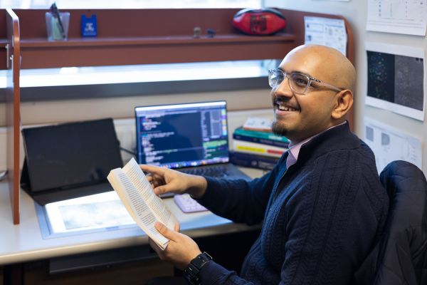 PhD student Dr Rishi Ramani sitting at a desk, holding a book and smiling at someone off camera