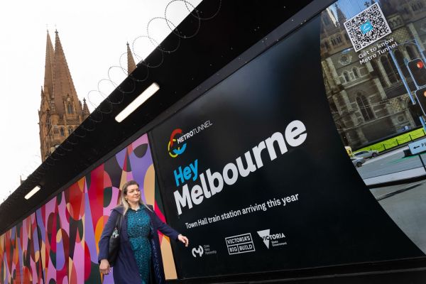 Associate Professor Rita Hardiman walks from left to right past hoardings that have the words: MetroTunnel written on them. The spire of St. Paul’s Cathedral can be seen from behind the hoardings in the upper left-hand corner. Credit: Peter Casamento