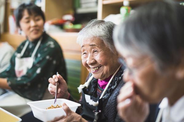 An elderly woman sits between two other women. She laughs while holding a container of food in her left hand and a pair of chopsticks in her right hand