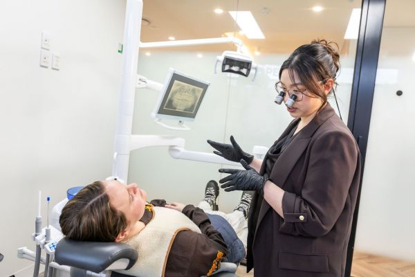 A patient lies in the dental chair at Luminaire Dental in Spotswood, looking up at Dr Jenny Dang who speaks to her while wearing black gloves. A monitor behind them shows an X-ray of teeth