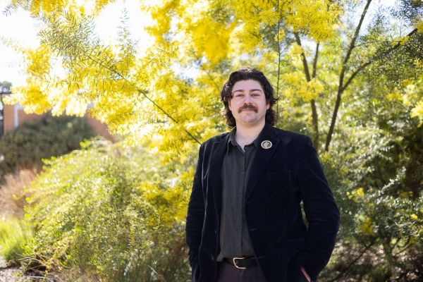 Portrait of Patrick Mercer, (alum) Wadawurrung man from Ballarat and Geelong and First Nations lecturer at the Melbourne Dental School, standing in front of a flowering wattle tree