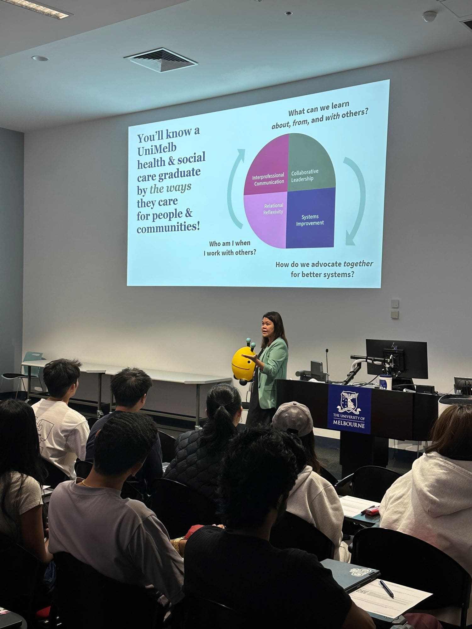 Vivienne Mak, standing in front of students, seated in rows, and delivering a PowerPoint presentation on teamwork to dentistry students
